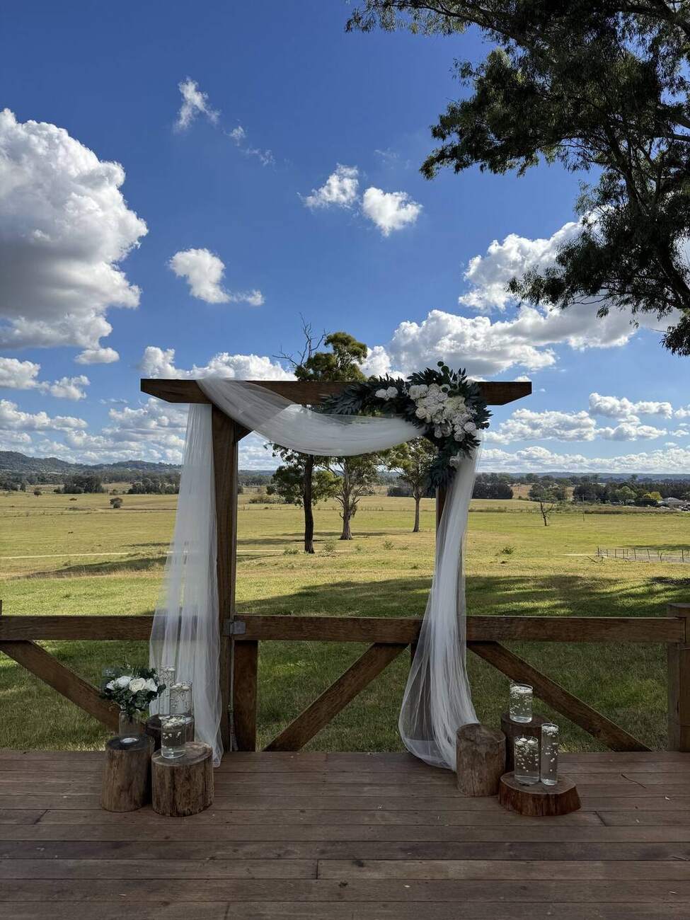 Rustic timber wedding arch with white drape and florals against a rural backdrop